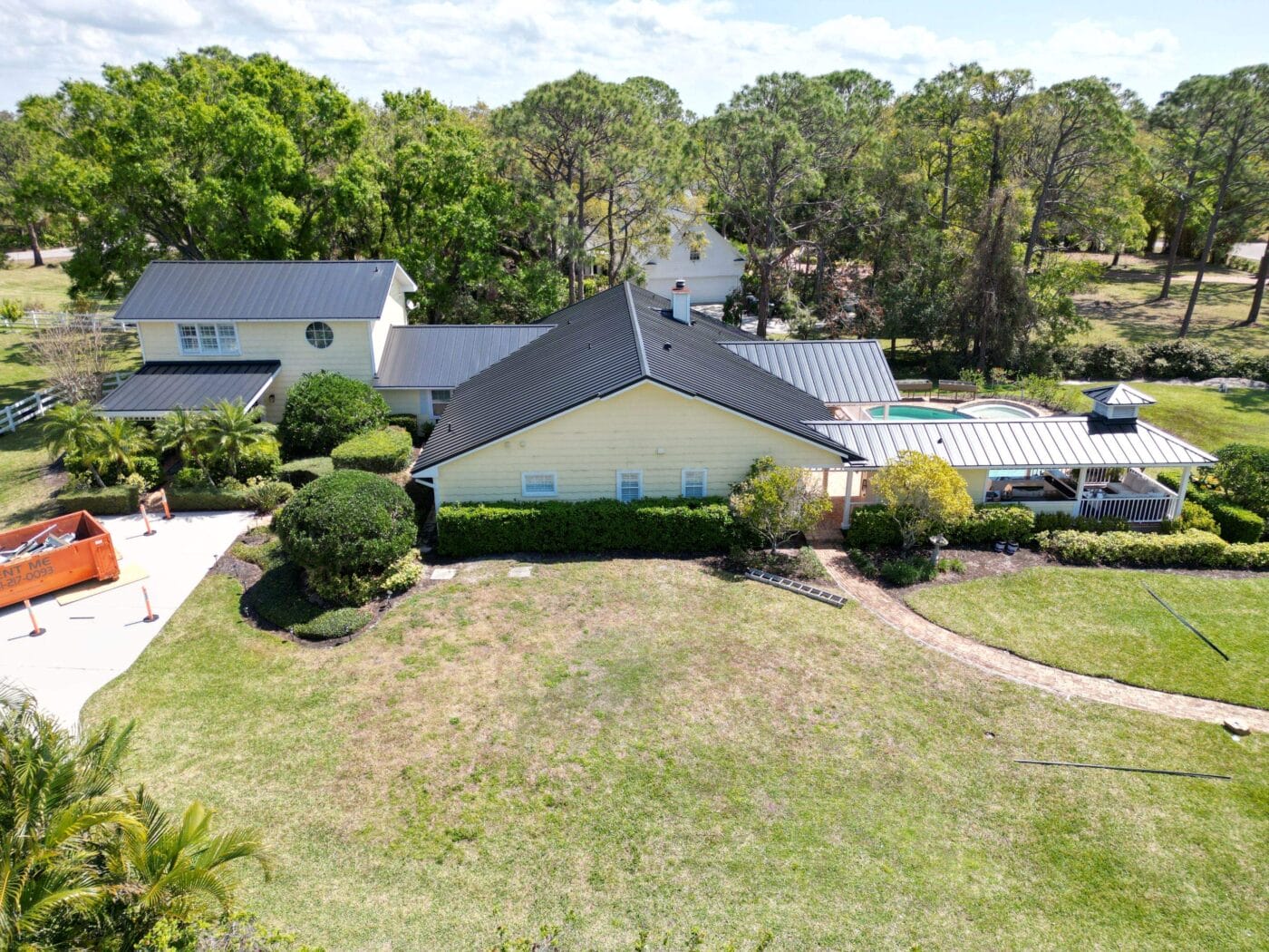 Aerial view of a large house with dark roofing and stylish siding, nestled among trees and a lush lawn. A swimming pool shimmers nearby, while a white structure and an orange dumpster rest by the driveway.