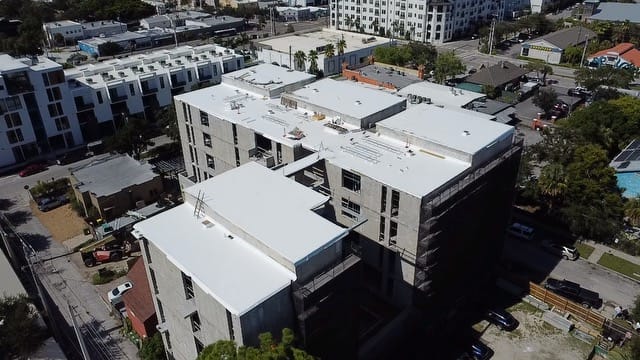 Aerial view of a multi-story building under construction by a General Contractor Sarasota FL, featuring a finished white roof and surrounded by other buildings and parked cars.
