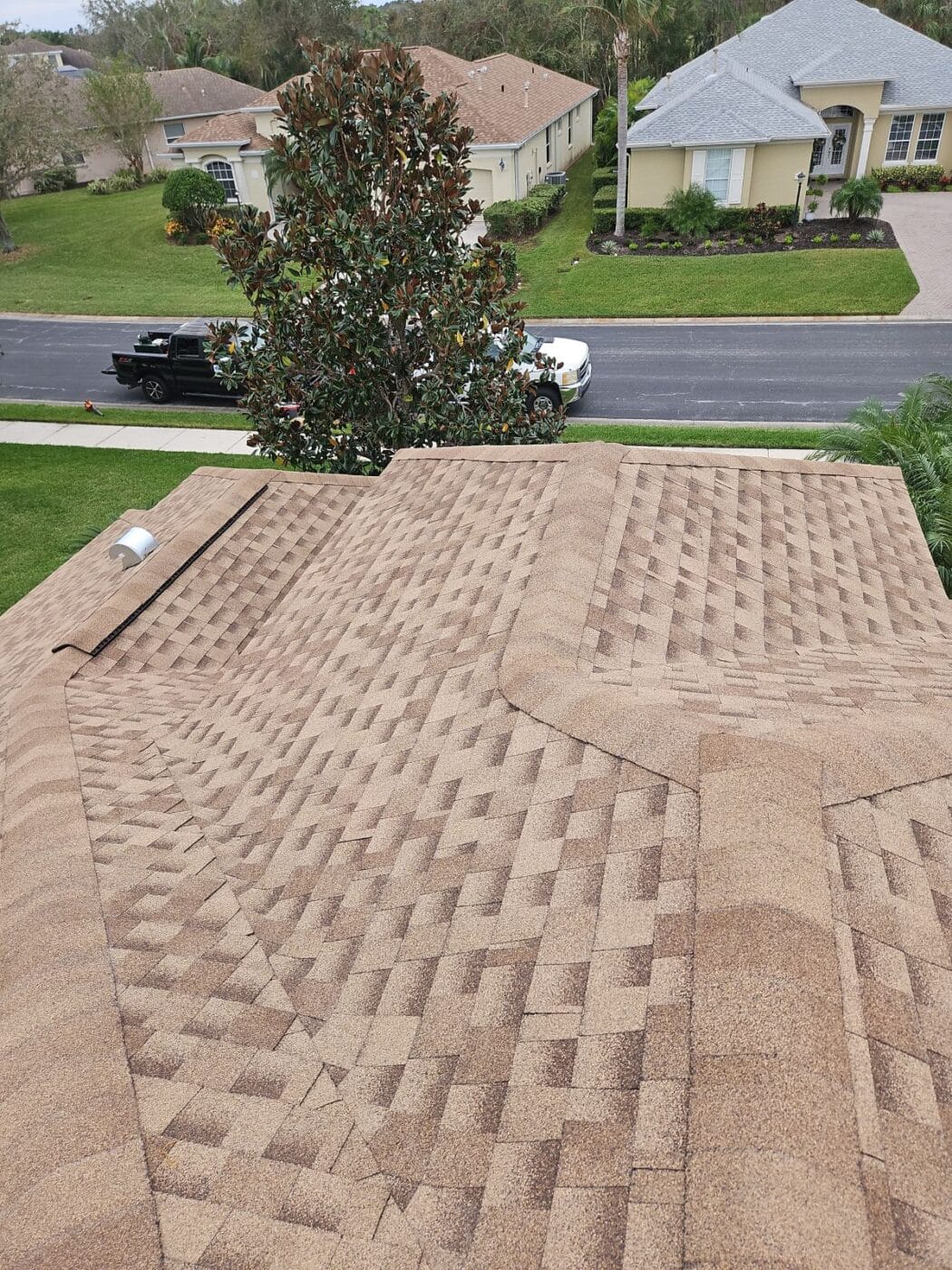 A view from a rooftop with brown asphalt shingles, showing intersecting rooflines; suburban houses, cars, and trees are visible in the background.
