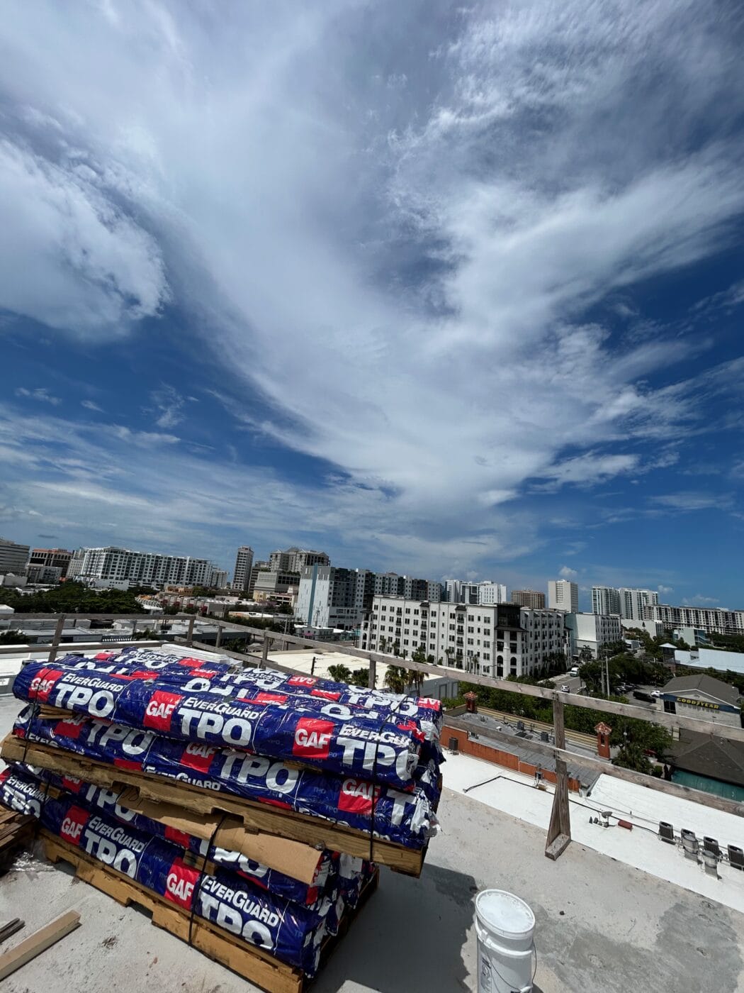 Stacks of TPO EverGuard roofing material on a rooftop overlook a cityscape with mid-rise buildings under a partly cloudy blue sky, ready for installation by a trusted General Contractor Sarasota FL.