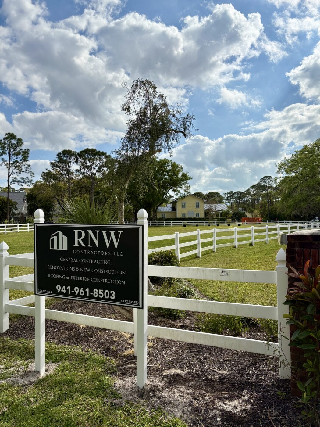 A white fenced yard with a green RNW Contractors LLC sign in the foreground highlights this yellow house, nestled among trees under a partly cloudy sky—your trusted General Contractor Sarasota FL.