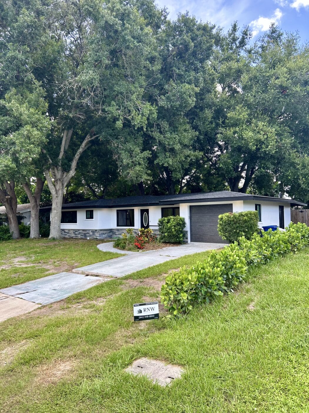 This single-story house boasts a pristine white facade and dark roof, complemented by elegant windows and doors. Nestled among trees and lush grass, it features a driveway leading to a black garage door with an 