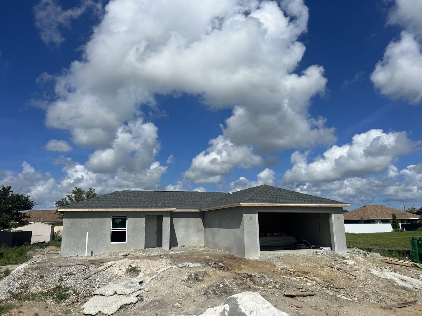 A single-story house under construction, featuring gray exterior walls and an open garage, is set against a blue sky with scattered clouds. Windows and doors await installation, while building materials are scattered on the ground in front.