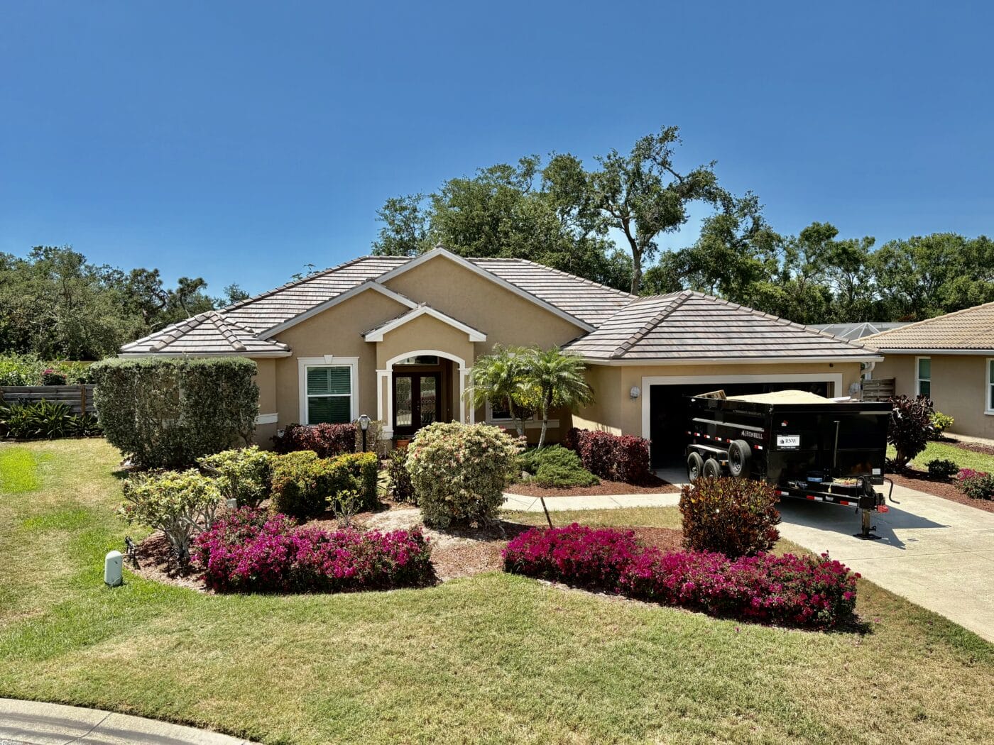 Single-story beige new construction house with a well-kept front yard, shrubs, and a trailer parked in the driveway under a clear blue sky.