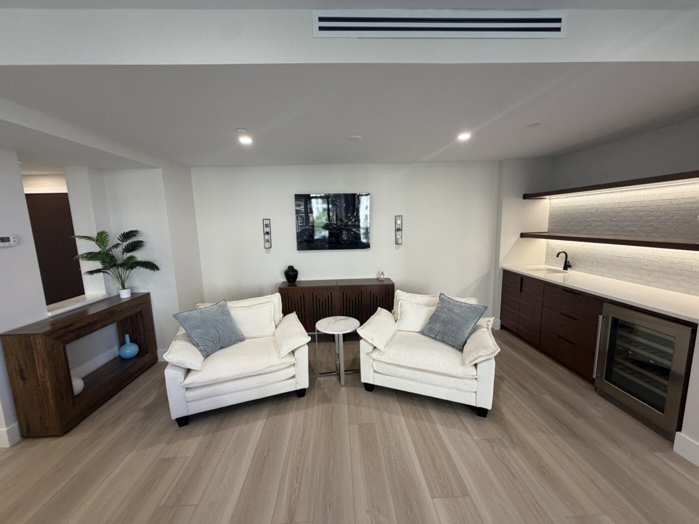 A modern living area, recently refreshed by a Whole Home Renovation, features two white armchairs, a small round table, wall-mounted TV, wood console, and a kitchenette with dark cabinets and a wine fridge.