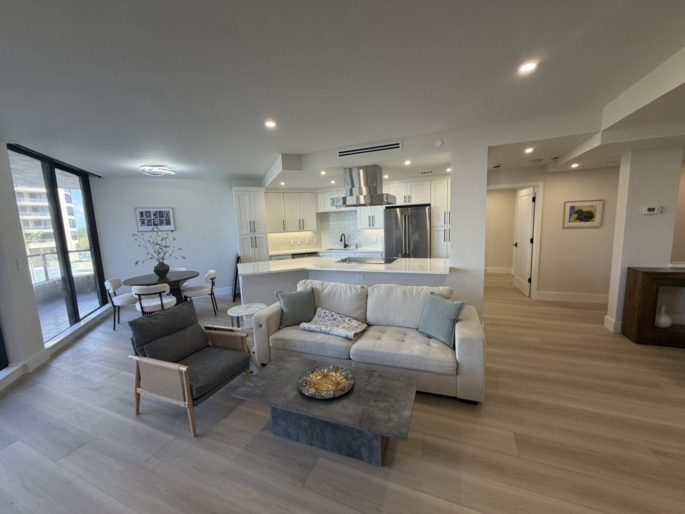 Open-plan living area with a sofa, armchair, coffee table, and dining table near glass doors; part of a recent Whole Home Renovation featuring a modern kitchen with white cabinets and stainless steel appliances in the background.