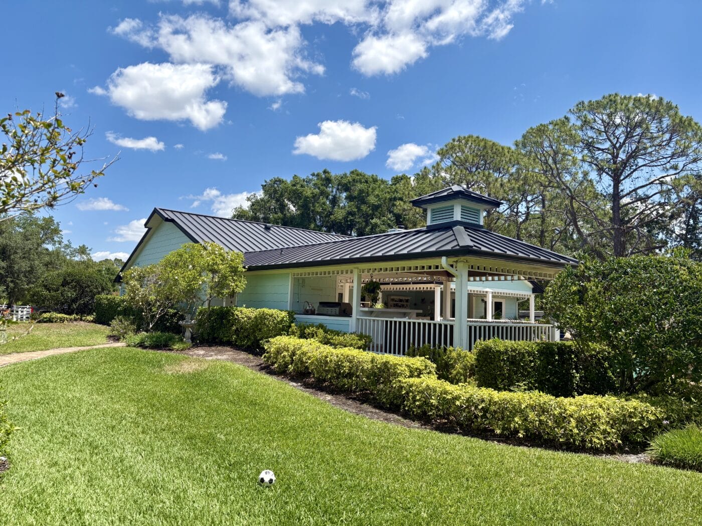 A light green new construction house with a covered porch sits amid neatly trimmed bushes and a grassy lawn under a blue sky with scattered clouds. A soccer ball lies on the grass.