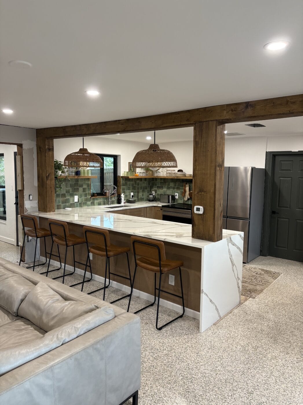 Modern kitchen with a marble island, four wooden barstools, green tiled backsplash, stainless steel appliances, and wooden pendant lights—all beautifully crafted by a General Contractor Sarasota FL—viewed from the living area.