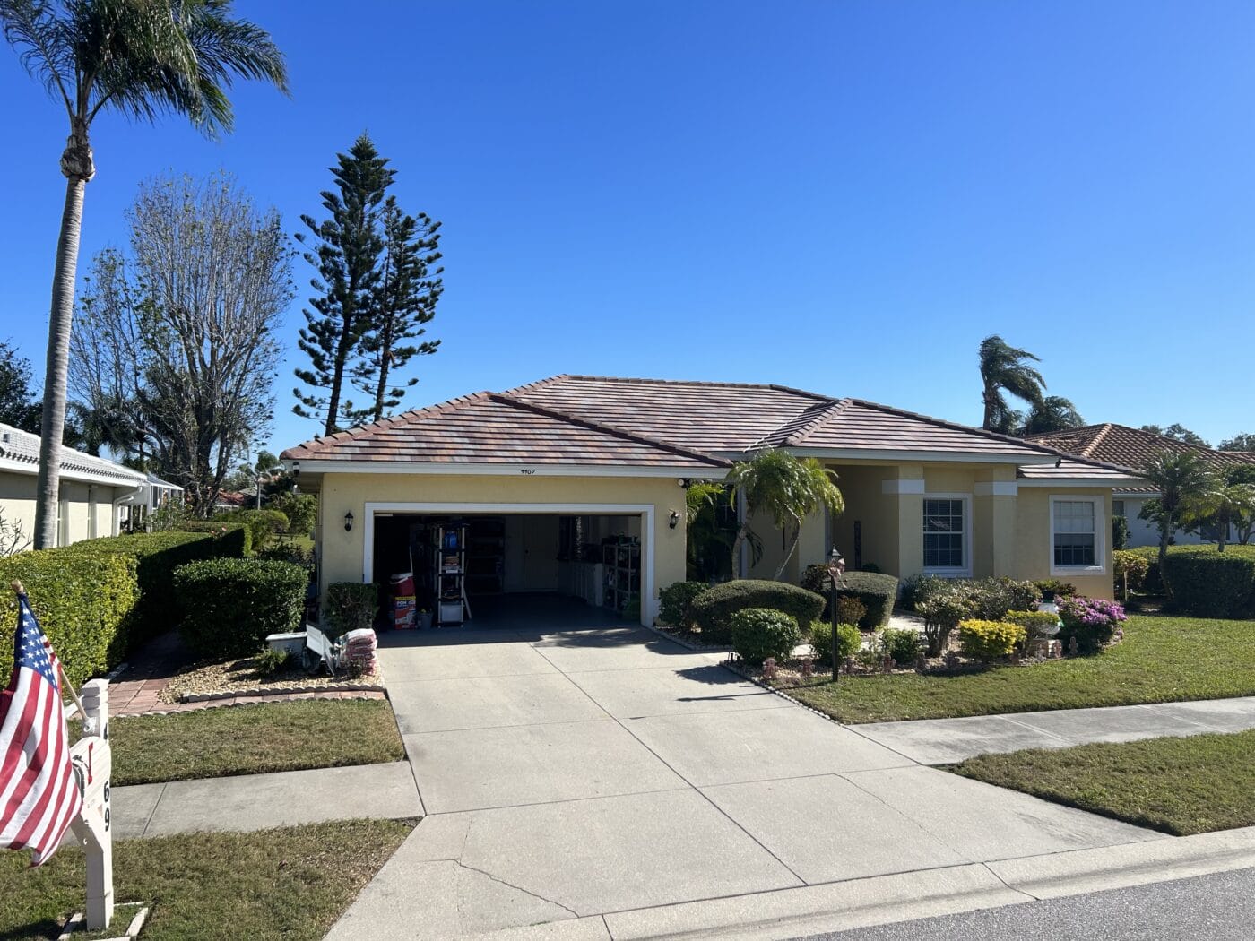 A single-story yellow house with a two-car garage, palm trees, and neatly trimmed shrubs in the front yard under a clear blue sky.