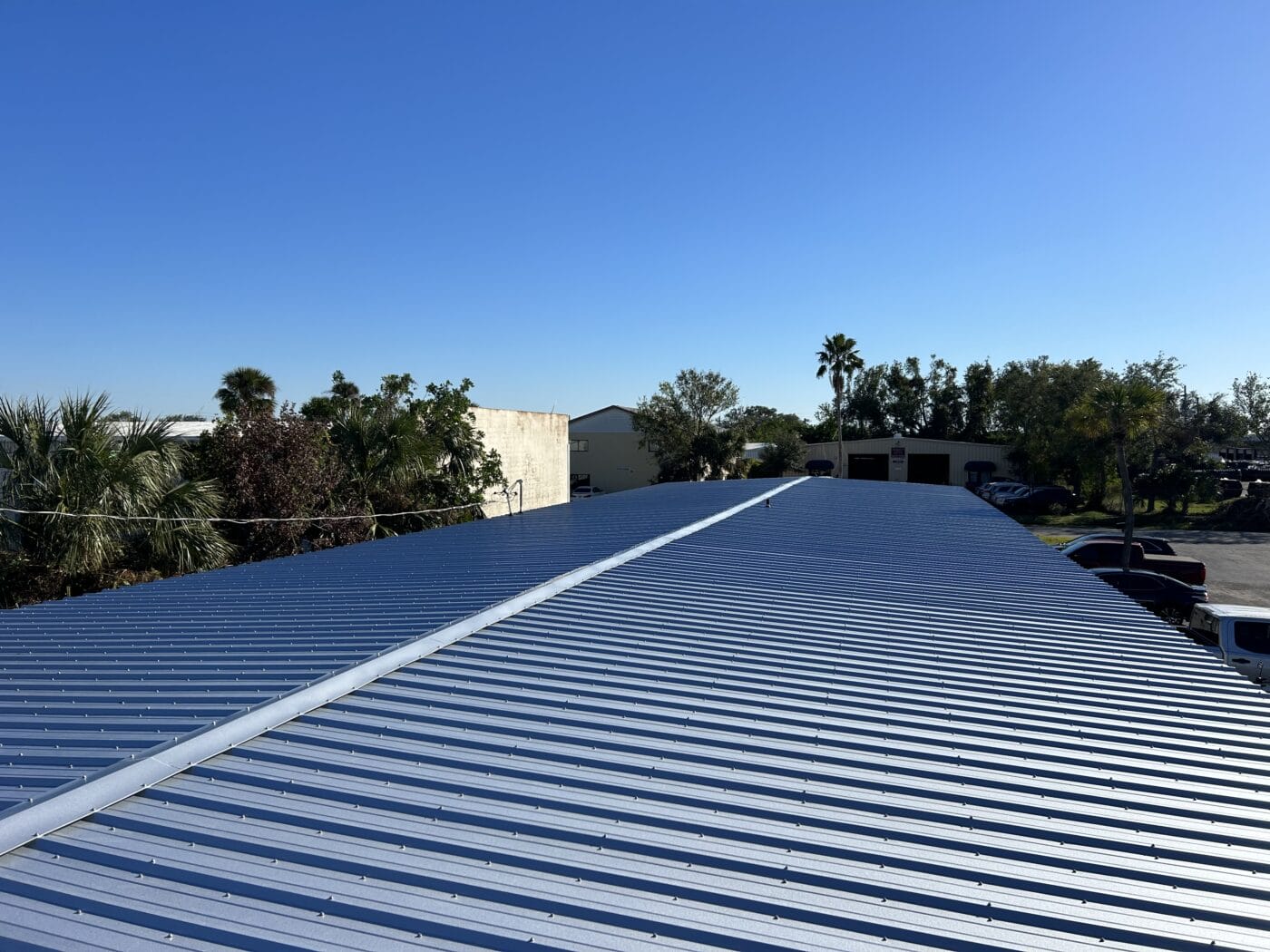 View of a large metal industrial roof beneath a clear blue sky, with trees on the left and buildings and parked vehicles in the background—an example of quality work by a General Contractor Sarasota FL.