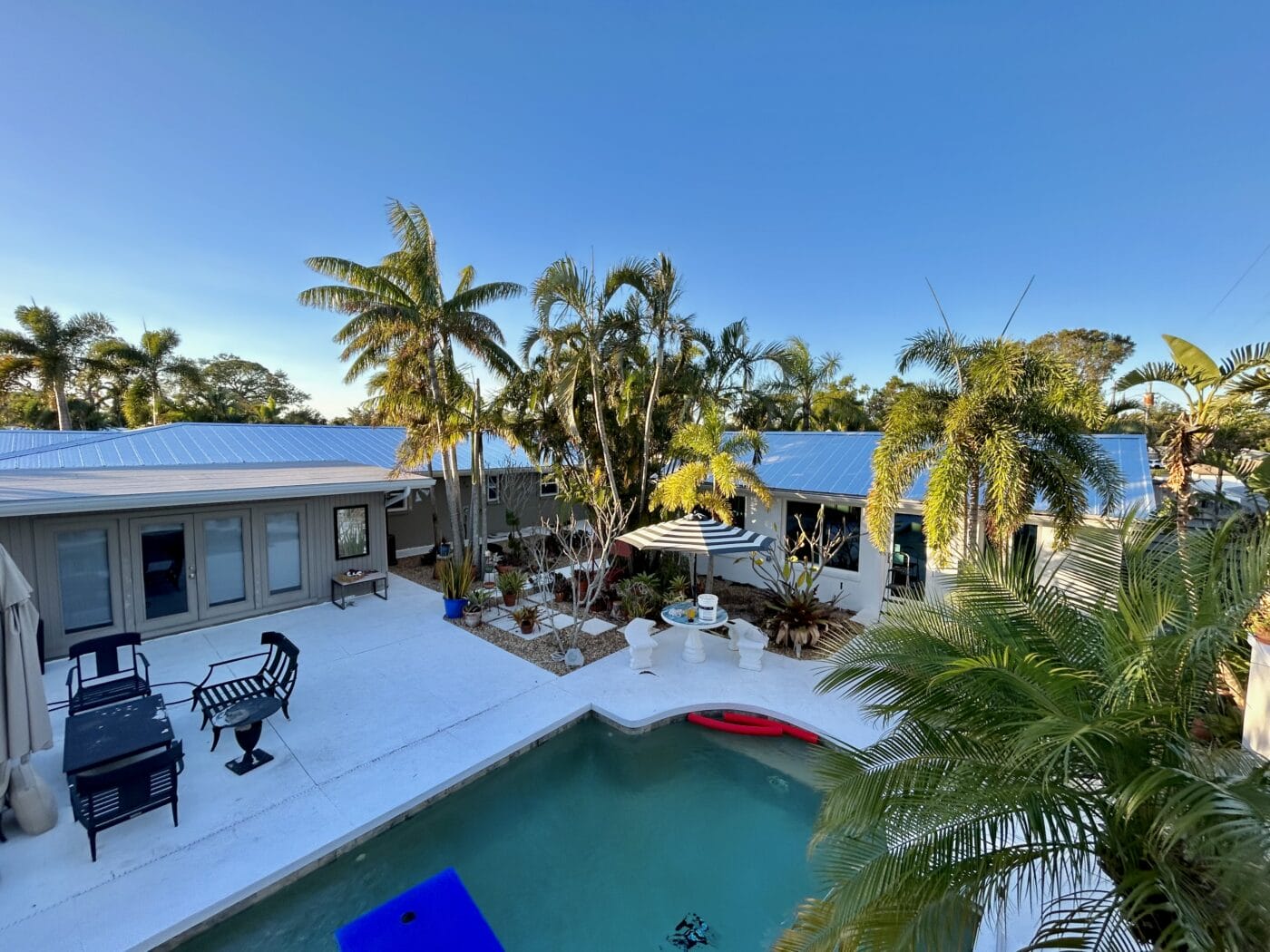 Outdoor pool area surrounded by tropical plants, lounge chairs, patio furniture, and two single-story buildings under a clear blue sky—expertly crafted by a trusted General Contractor Sarasota FL.