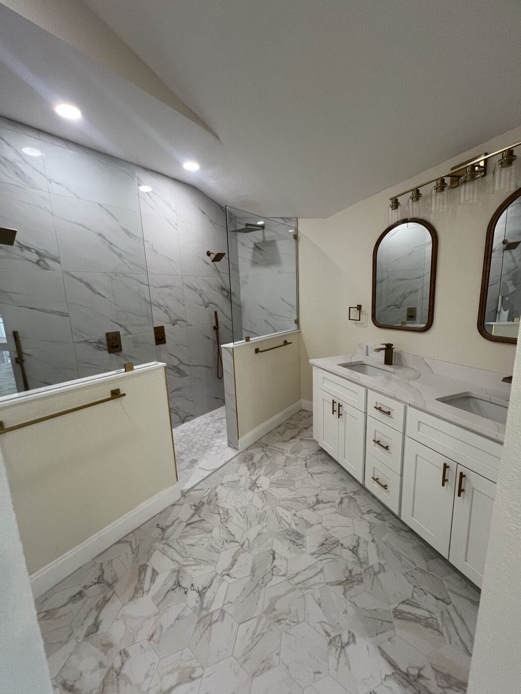 Modern bathroom remodeling with white and gray marble surfaces, double sinks, two mirrors, gold fixtures, and a walk-in shower with glass partition.