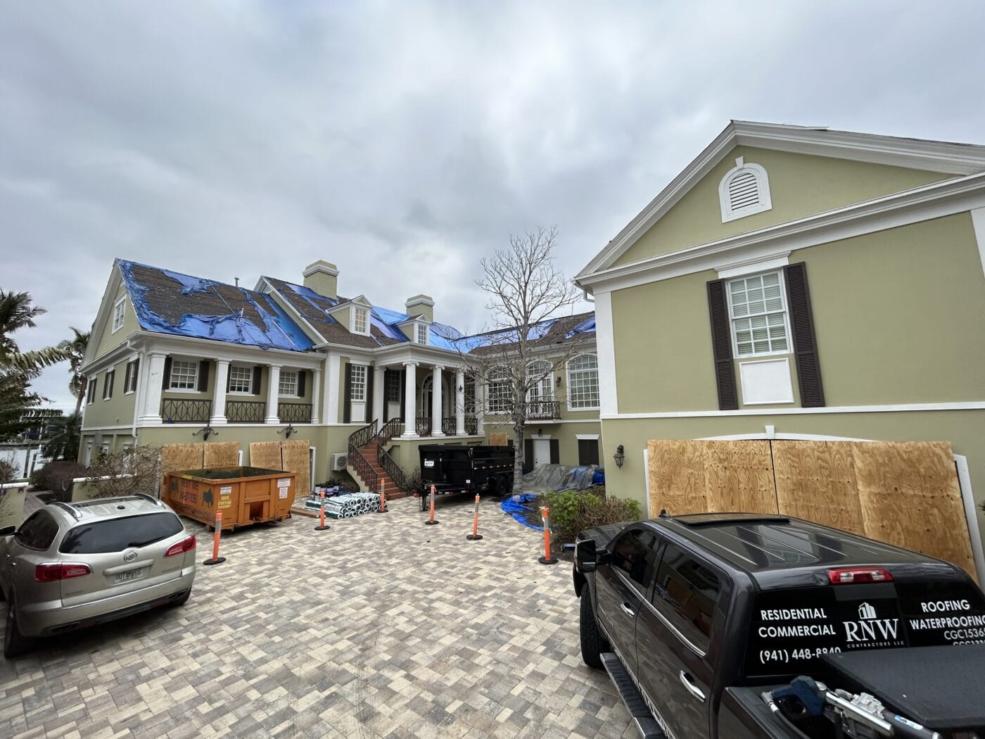 A large house with tarps on the roof, construction materials, a dumpster, and work vehicles parked in the driveway under a cloudy sky.