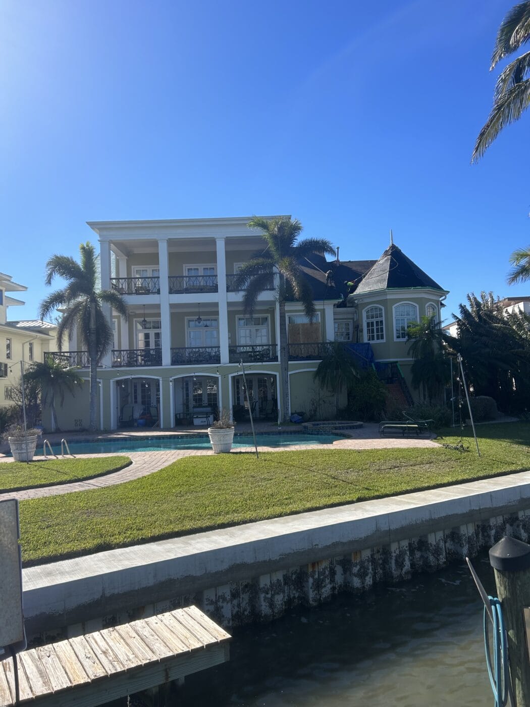Large waterfront house with three stories, featuring expansive windows and elegant doors leading to multiple balconies. Surrounded by palm trees, a curved pool and grassy lawn beckon in the foreground, under a clear blue sky.