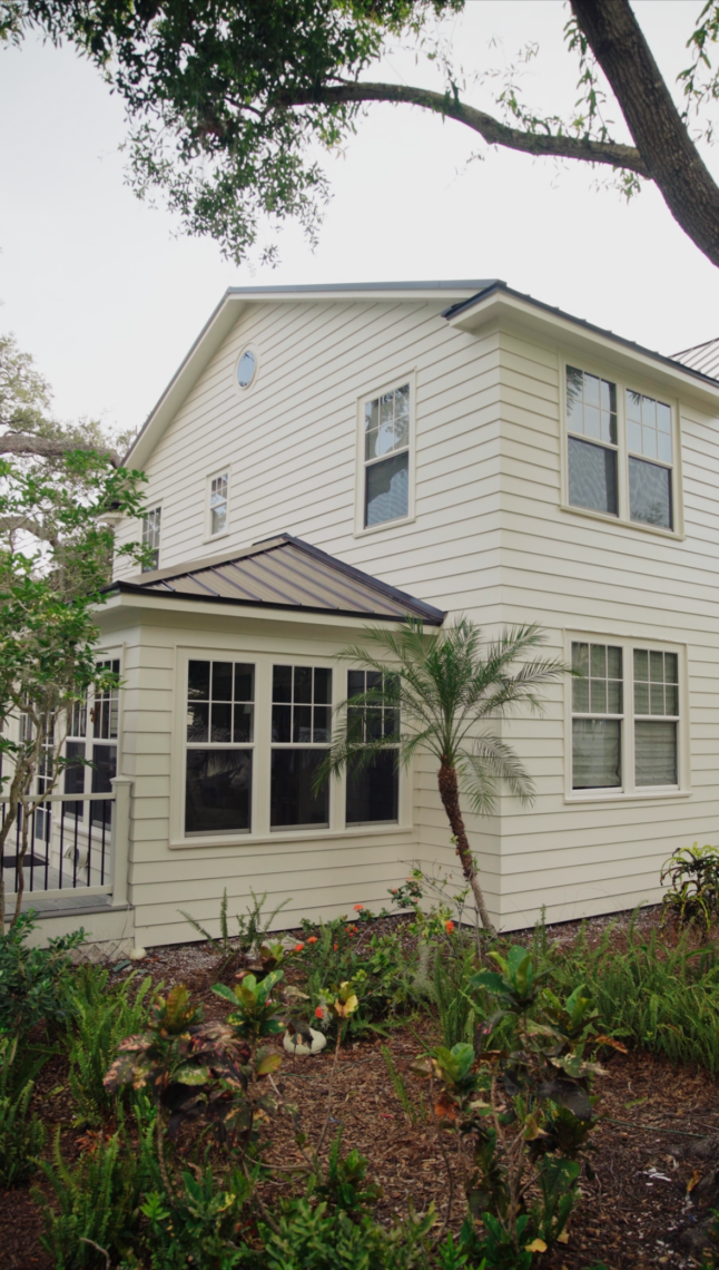 Two-story white house with large windows and stylish doors, a small porch, and surrounding greenery including a palm tree and various plants.