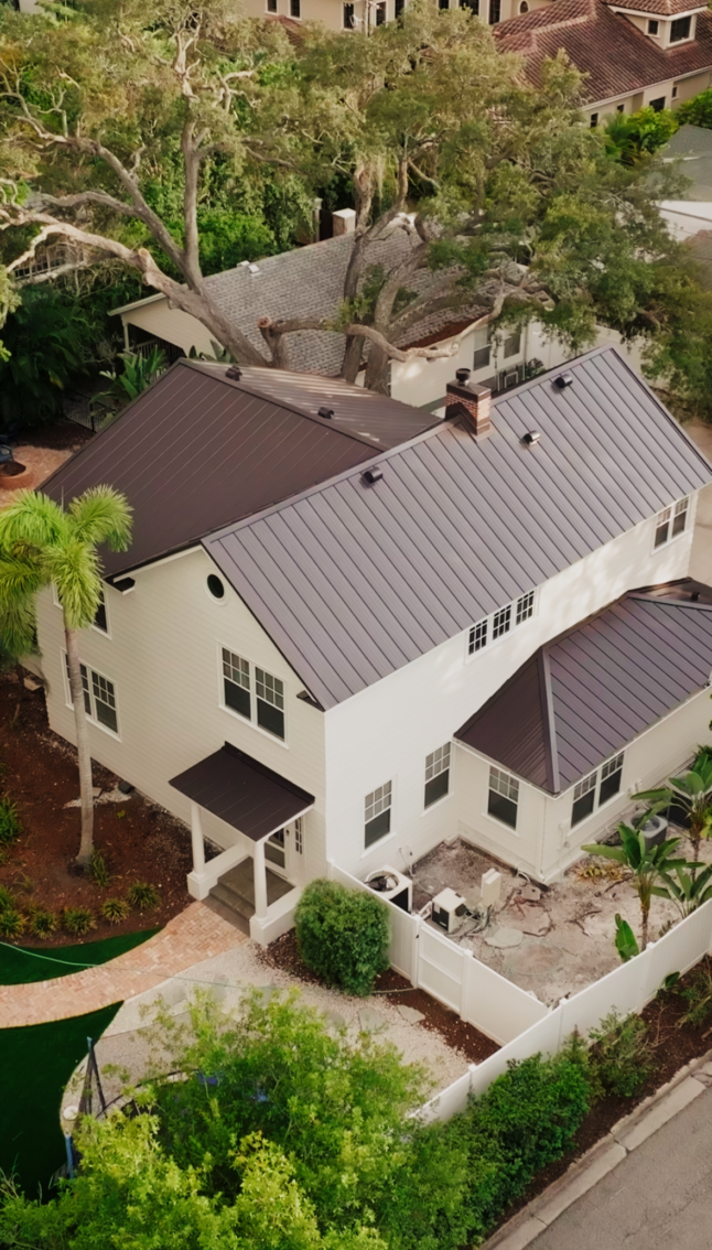 Aerial view of a two-story white house with sleek siding, a dark metal roof, a small fenced patio, and surrounding greenery and trees.