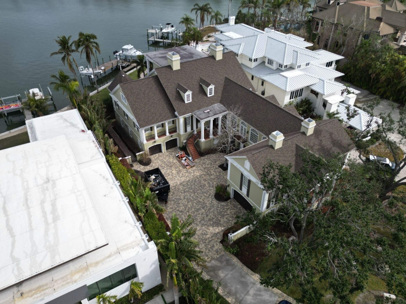 A large waterfront house with a brown roof, multiple chimneys, and a paved driveway, surrounded by palm trees and neighboring homes along a canal.