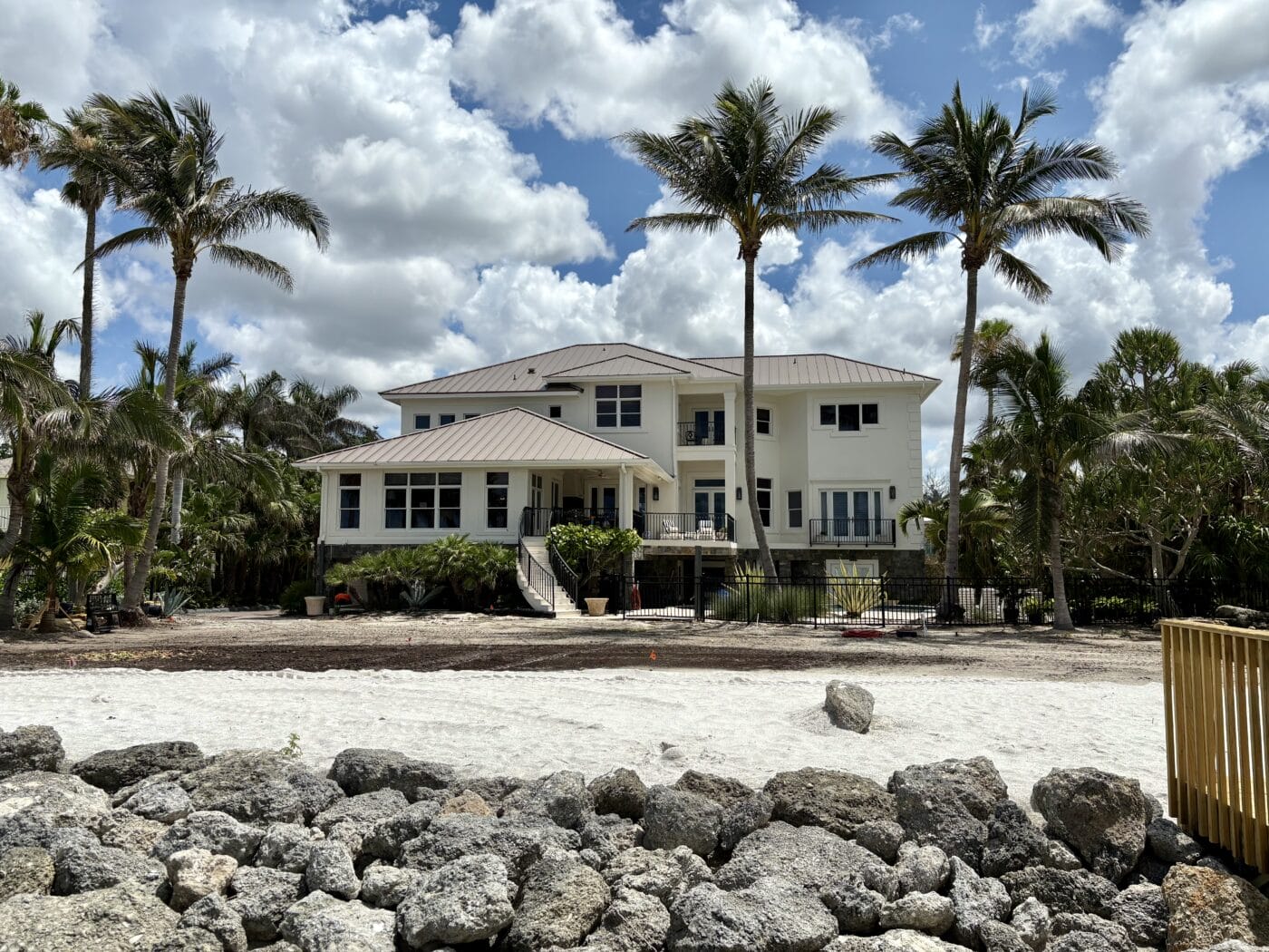 A large white two-story house with a metal roof is surrounded by palm trees, set behind a sandy area and a line of rocks under a partly cloudy sky.