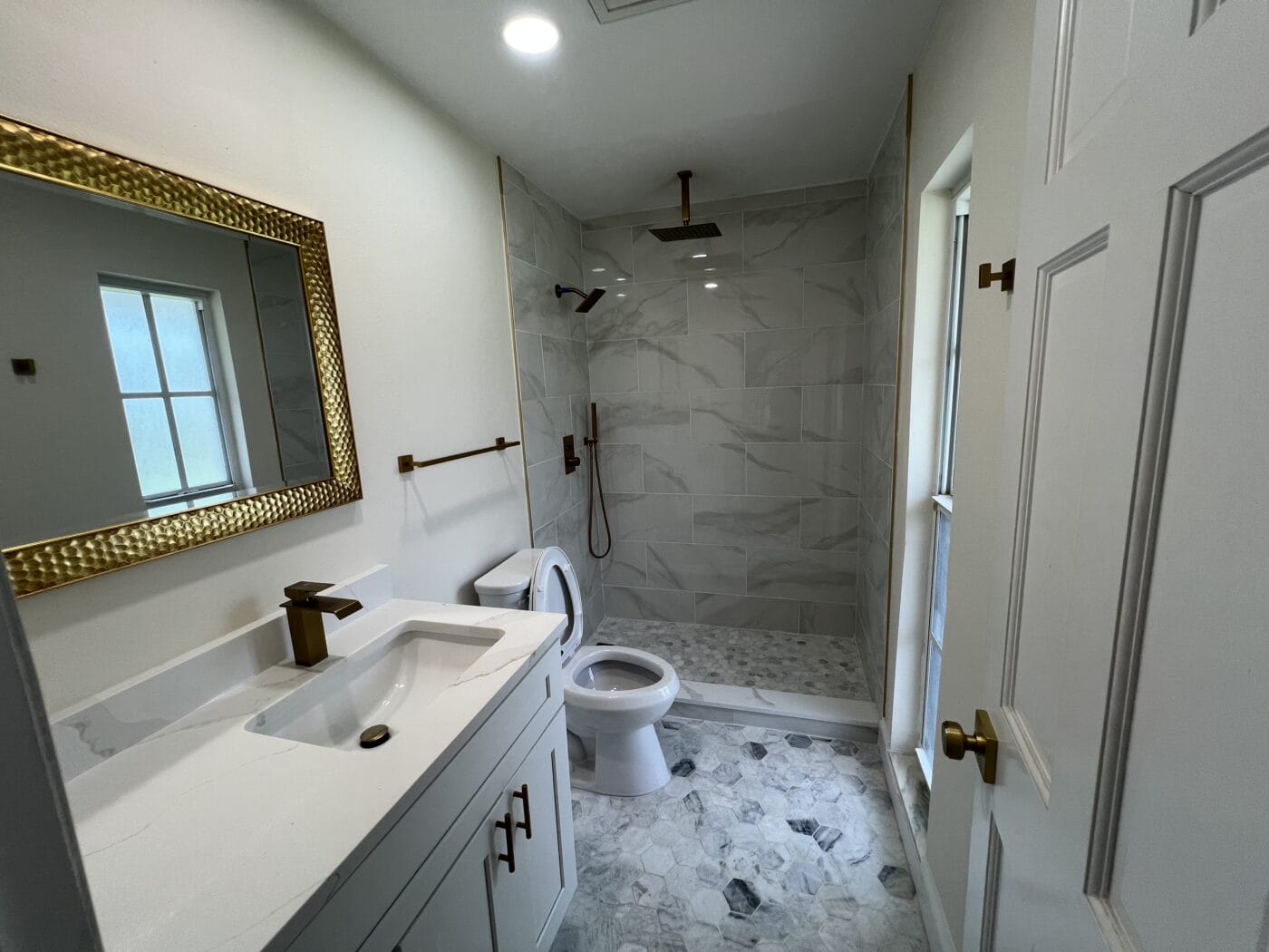 Modern bathroom with a white vanity, gold accents, a wall-mounted mirror, toilet, and a walk-in shower with marble tile.
