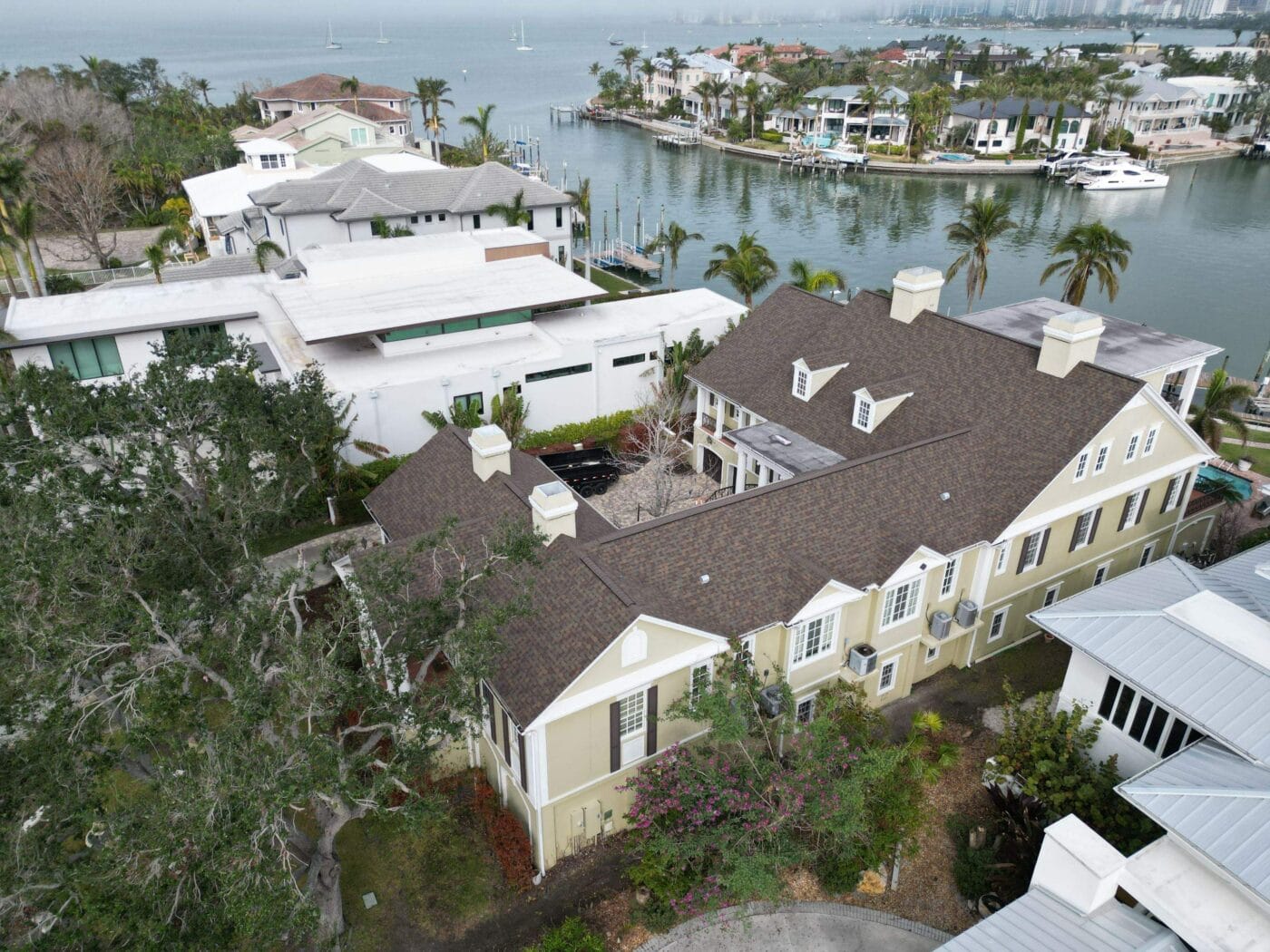 Aerial view of large waterfront homes with landscaped yards, palm trees, and docks along a calm bay with boats and nearby houses.